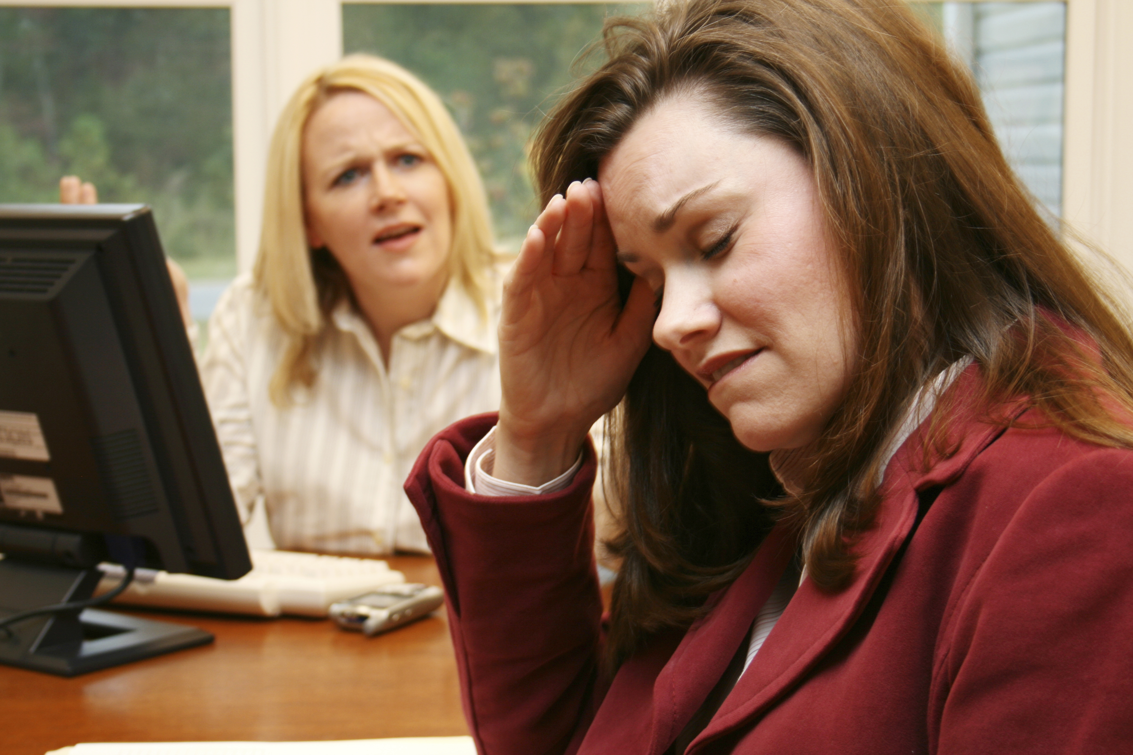 two women in an office setting, one in the background angrily speaking to the one in the foreground, who is holding her forehead as if she has a headache