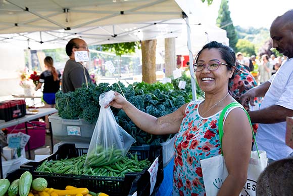 Photograph of a woman holding a bag of produce at a farmers market stall.