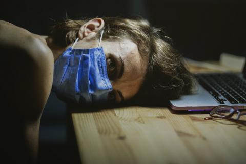 Photograph of a person wearing a medical mask resting their head on a desk in front of a computer.