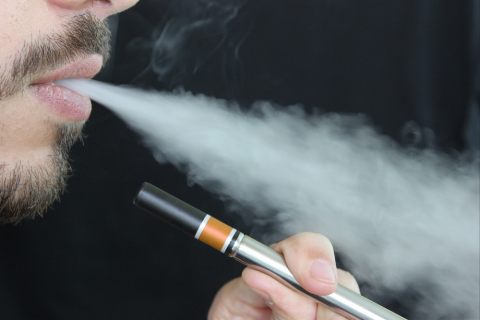 Photo of the lower quarter of a man's face blowing out smoke, while he holds a vape pen.