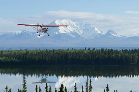 Photograph of small airplane flying over a lake with mountains in the background.