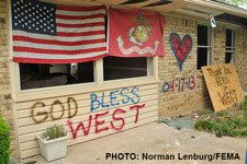 Photograph of a boarded up house with an American flag and "God Bless West" painted on it.