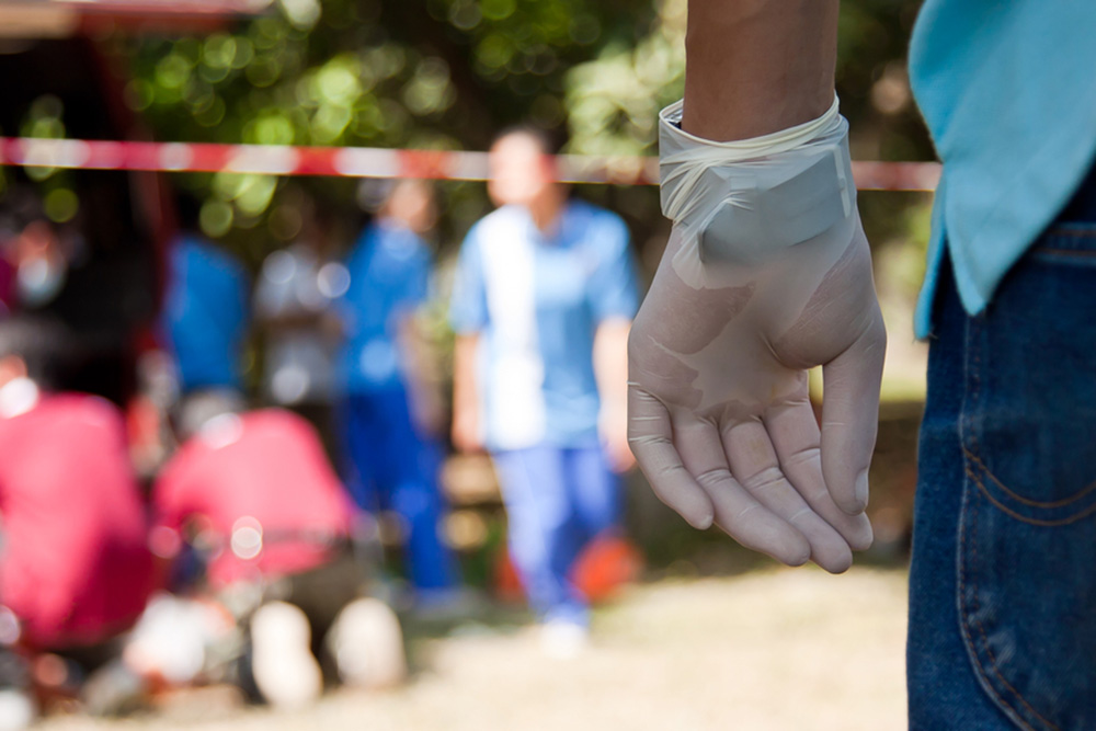 Photograph of a man's gloved hand with EMTs working in the background.